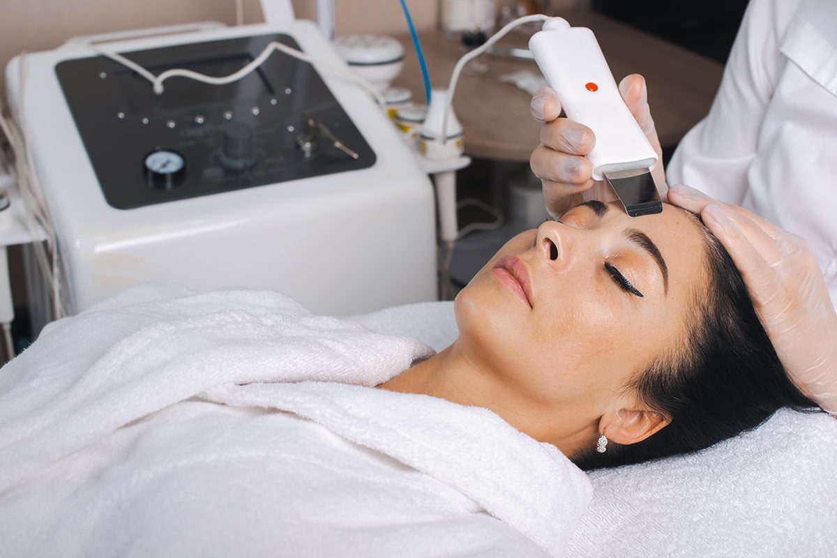 Cosmetologist's Hands In Gloves Doing An Ultrasound Cleaning Of A Caucasian Woman's Face. Beautiful Portrait. Cleaning Machine. Cleaning Concept. Skin Care Concept. Rejuvenation Treatment.